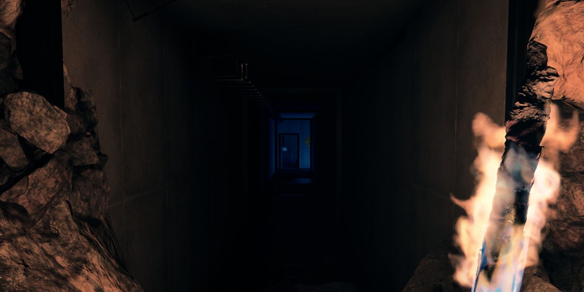 Una captura de pantalla de Sons of the Forest que muestra al personaje del jugador sosteniendo una antorcha encendida mientras mira hacia un pasillo largo y oscuro al final de una cueva con luz azul y una puerta al final.